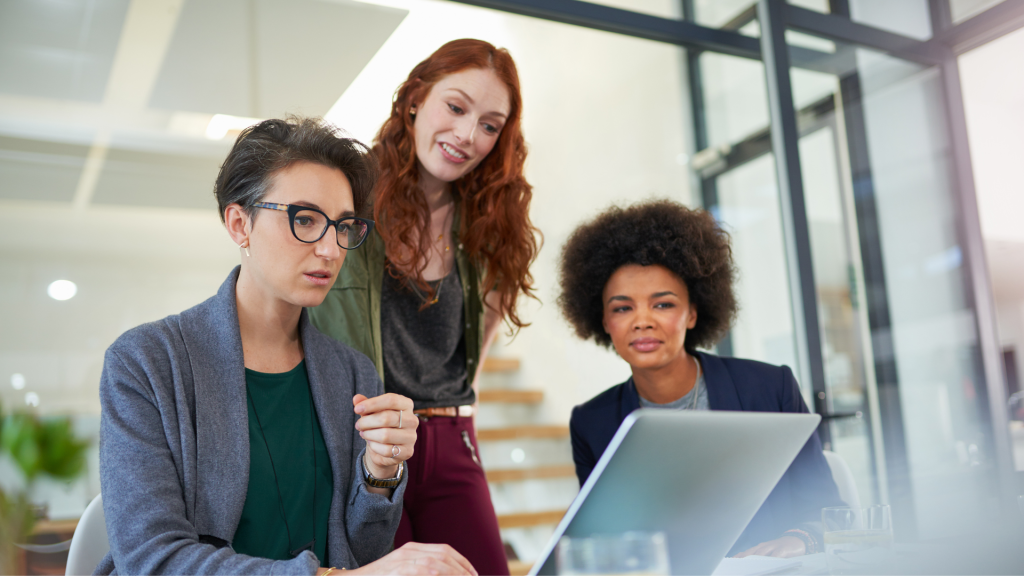 Three professional women in business attire gathered around a laptop, discussing work.
